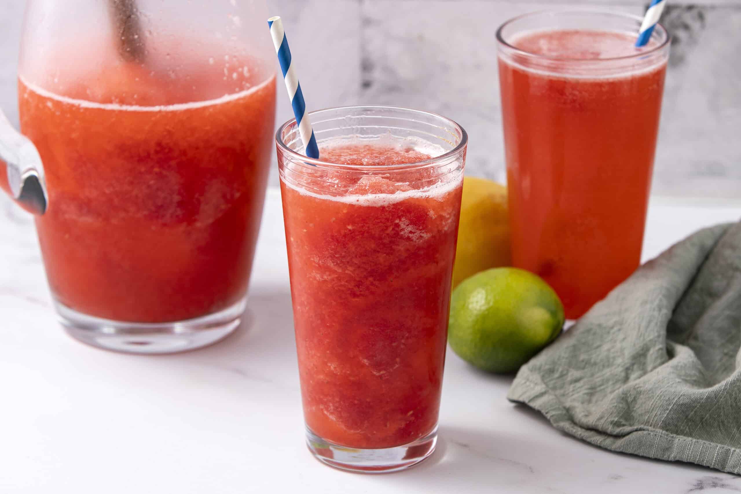 Refreshing watermelon lime juice served in tall glasses with blue and white striped straws, surrounded by fresh limes and a lemon, on a white marble surface with a rustic gray background.
