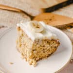Moist banana cake slice with creamy frosting and banana slices on top, served on a delicate white plate, highlighting delicious homemade banana dessert.