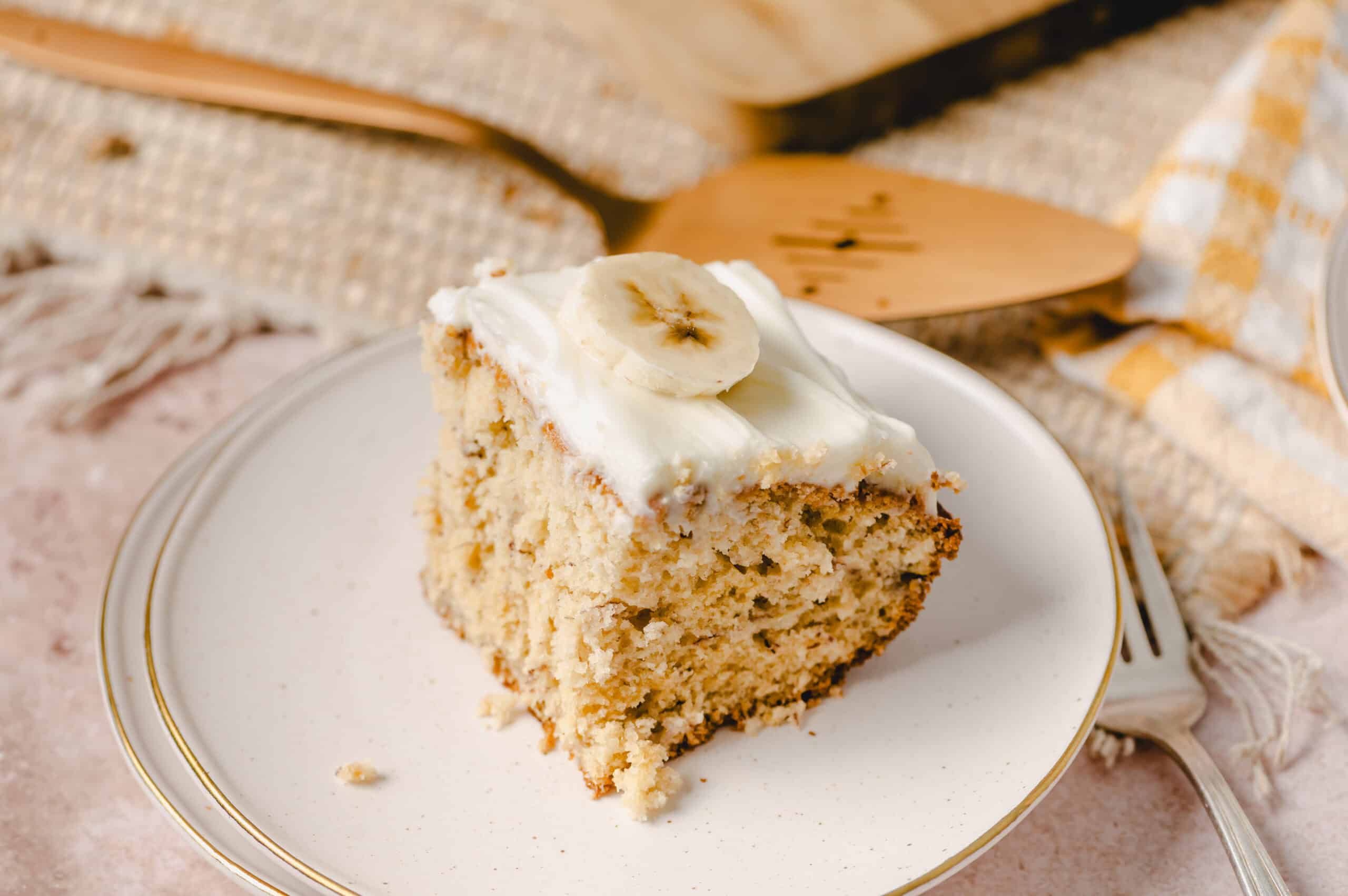 Moist banana cake slice with creamy frosting and banana slices on top, served on a delicate white plate, highlighting delicious homemade banana dessert.