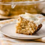 Moist homemade banana bread slice with creamy frosting on a white plate. Freshly baked banana bread topped with whipped cream, served on a rustic table with a yellow and white striped cloth.