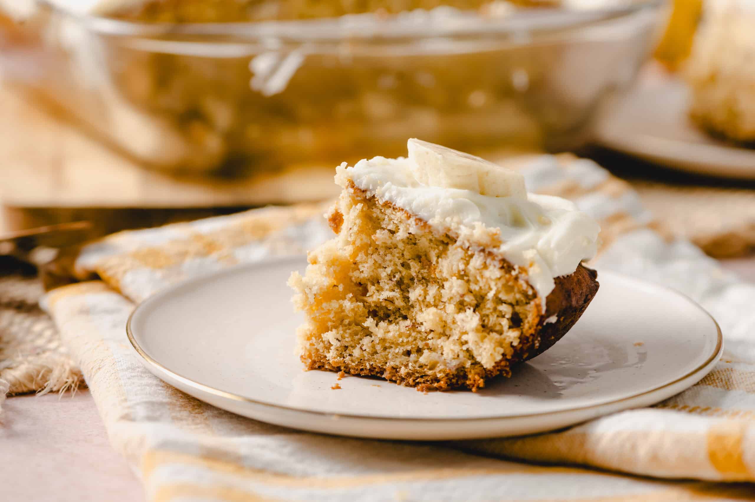 Moist homemade banana bread slice with creamy frosting on a white plate. Freshly baked banana bread topped with whipped cream, served on a rustic table with a yellow and white striped cloth.