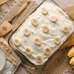 Creamy banana dessert with fresh banana slices on top, served in a glass baking dish, surrounded by bananas, a mixer spatula, plates, and a fork, perfect for easy and delicious banana pudding recipes.