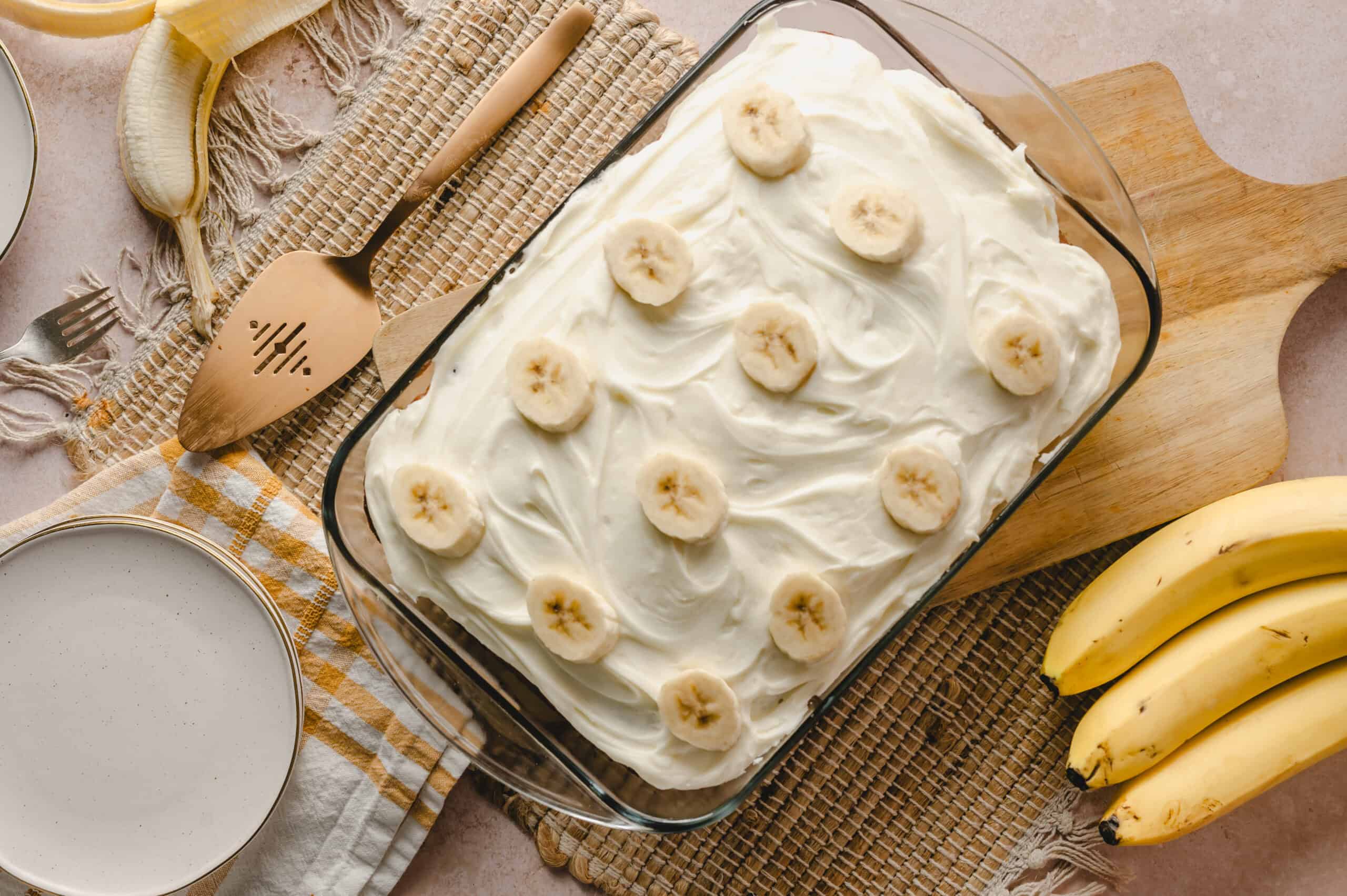 Creamy banana dessert with fresh banana slices on top, served in a glass baking dish, surrounded by bananas, a mixer spatula, plates, and a fork, perfect for easy and delicious banana pudding recipes.
