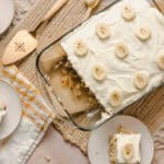Creamy banana pudding dessert with whipped topping and sliced bananas, served in a glass dish on a rustic table setting with bananas in the background.