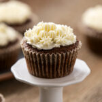 Delicious chocolate cupcake topped with creamy white frosting and decorative piped icing, served on a white cake stand with blurred cupcakes in the background.