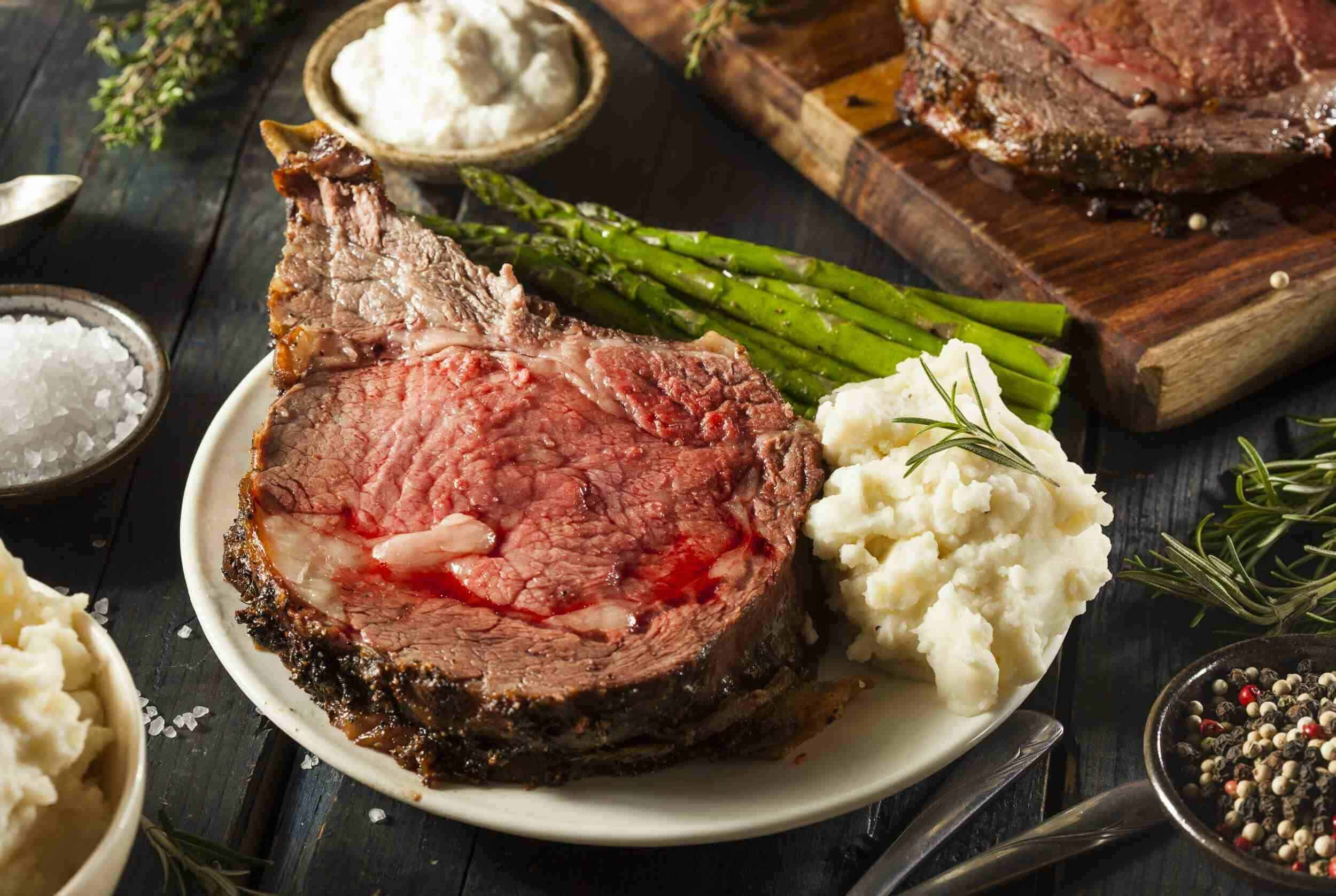 Juicy prime rib roast with garlic herb crust served with mashed potatoes, asparagus, and side dishes on rustic dark wooden table.