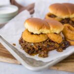 Savory sloppy joe sliders with onion rings on a white tray, served with dipping sauce, ready to enjoy classic comfort food.