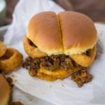 Juicy cheeseburger with crispy onion rings on white paper, melted cheese, seasoned ground beef patty, and soft sandwich bun, served with dipping sauces on the side.
