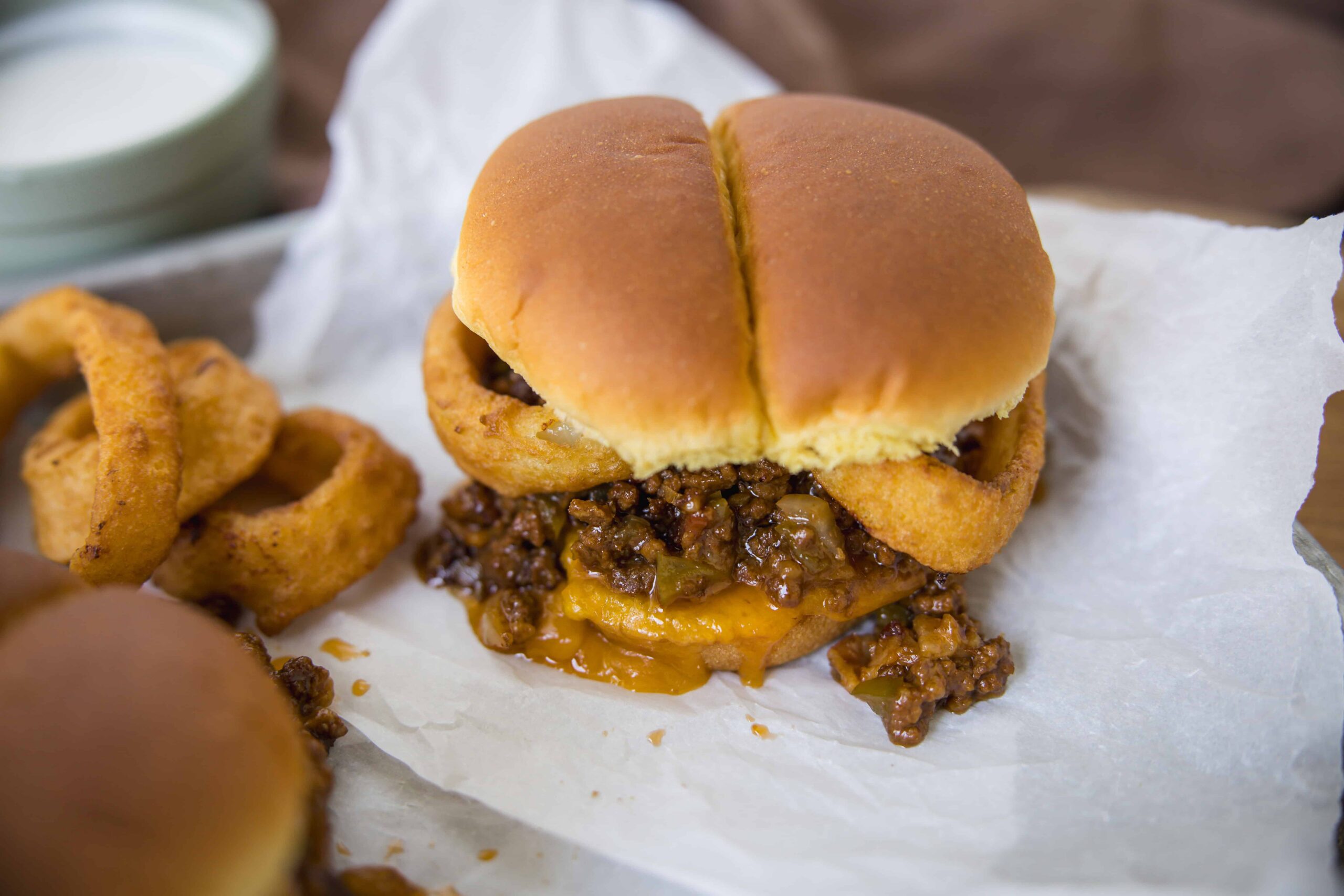 Juicy cheeseburger with crispy onion rings on white paper, melted cheese, seasoned ground beef patty, and soft sandwich bun, served with dipping sauces on the side.