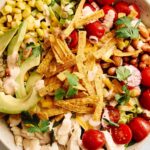 Fresh vegetable taco salad with cherry tomatoes, avocado, corn, beans, shredded chicken, crispy tortilla strips, and cilantro in a white bowl. Perfect healthy meal with colorful ingredients.