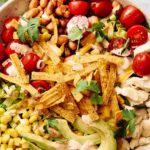 Colorful taco salad with fresh cherry tomatoes, grilled chicken, shredded cheese, avocado slices, corn, crispy tortilla strips, and cilantro in a white ceramic bowl.