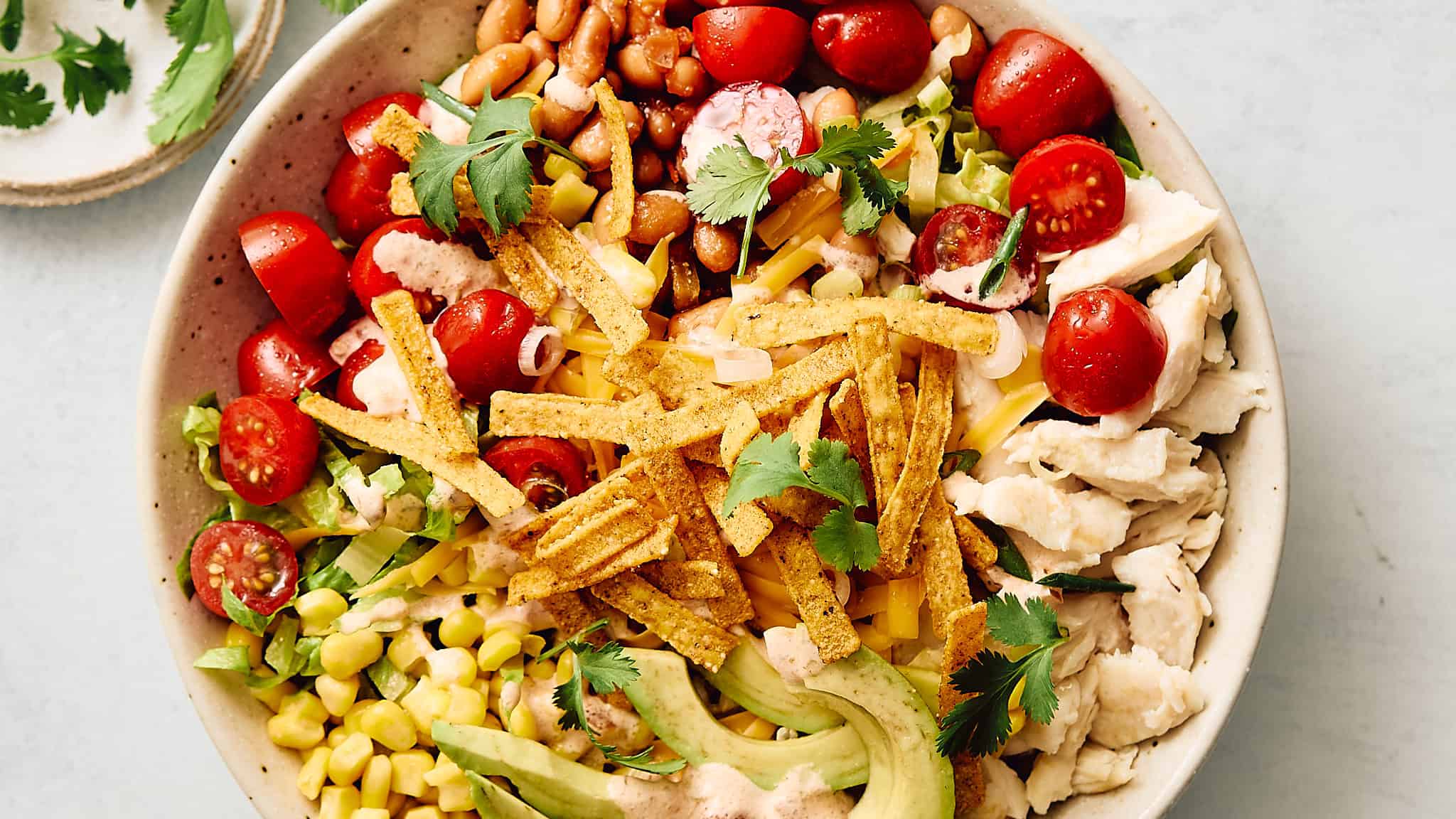 Colorful taco salad with fresh cherry tomatoes, grilled chicken, shredded cheese, avocado slices, corn, crispy tortilla strips, and cilantro in a white ceramic bowl.