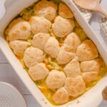 Golden baked chicken pot pie with flaky biscuit topping in a white casserole dish, served with fresh herbs, on a rustic white wooden table.