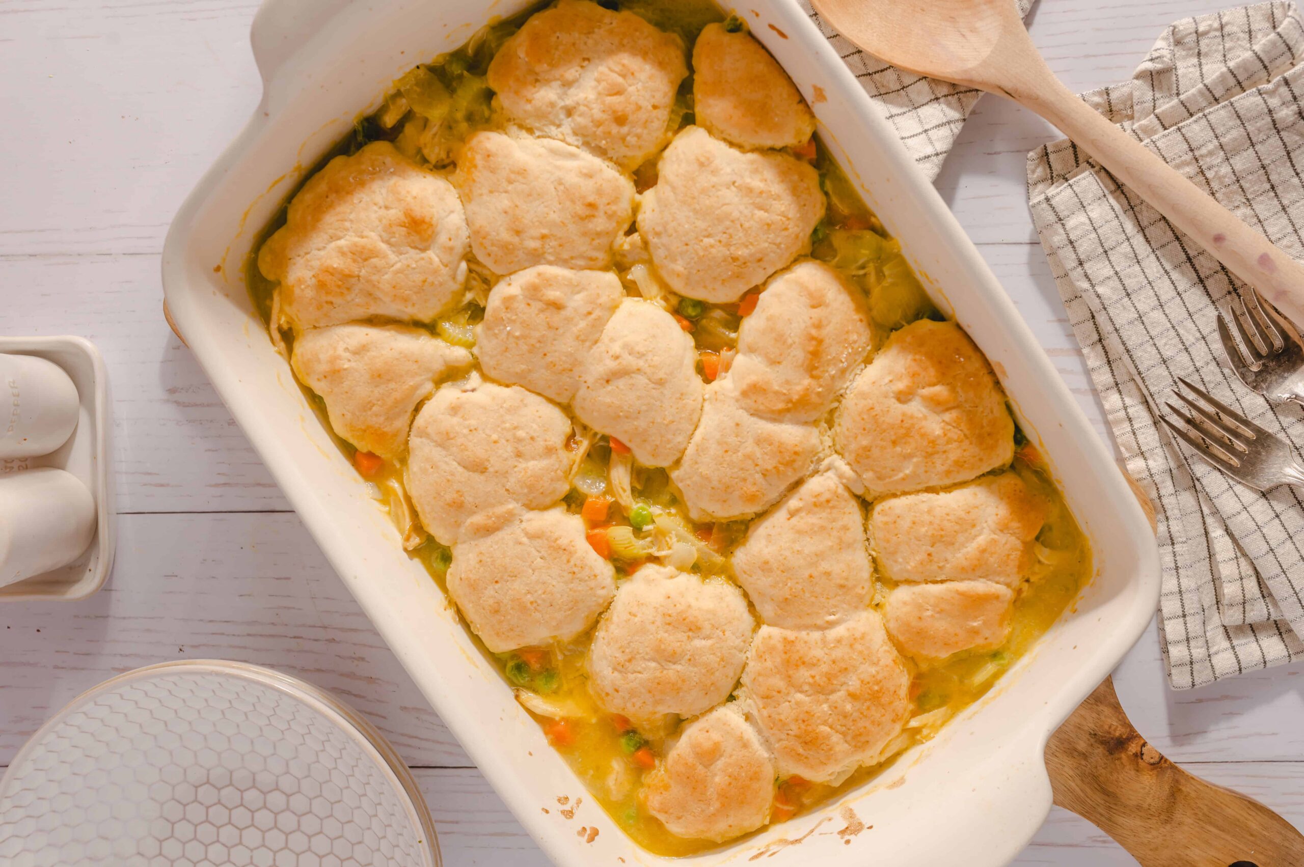 Golden baked chicken pot pie with flaky biscuit topping in a white casserole dish, served with fresh herbs, on a rustic white wooden table.