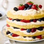 Fresh berry layered sponge cake with whipped cream and assorted berries on top and in between layers, served on a decorative plate for a delightful dessert.