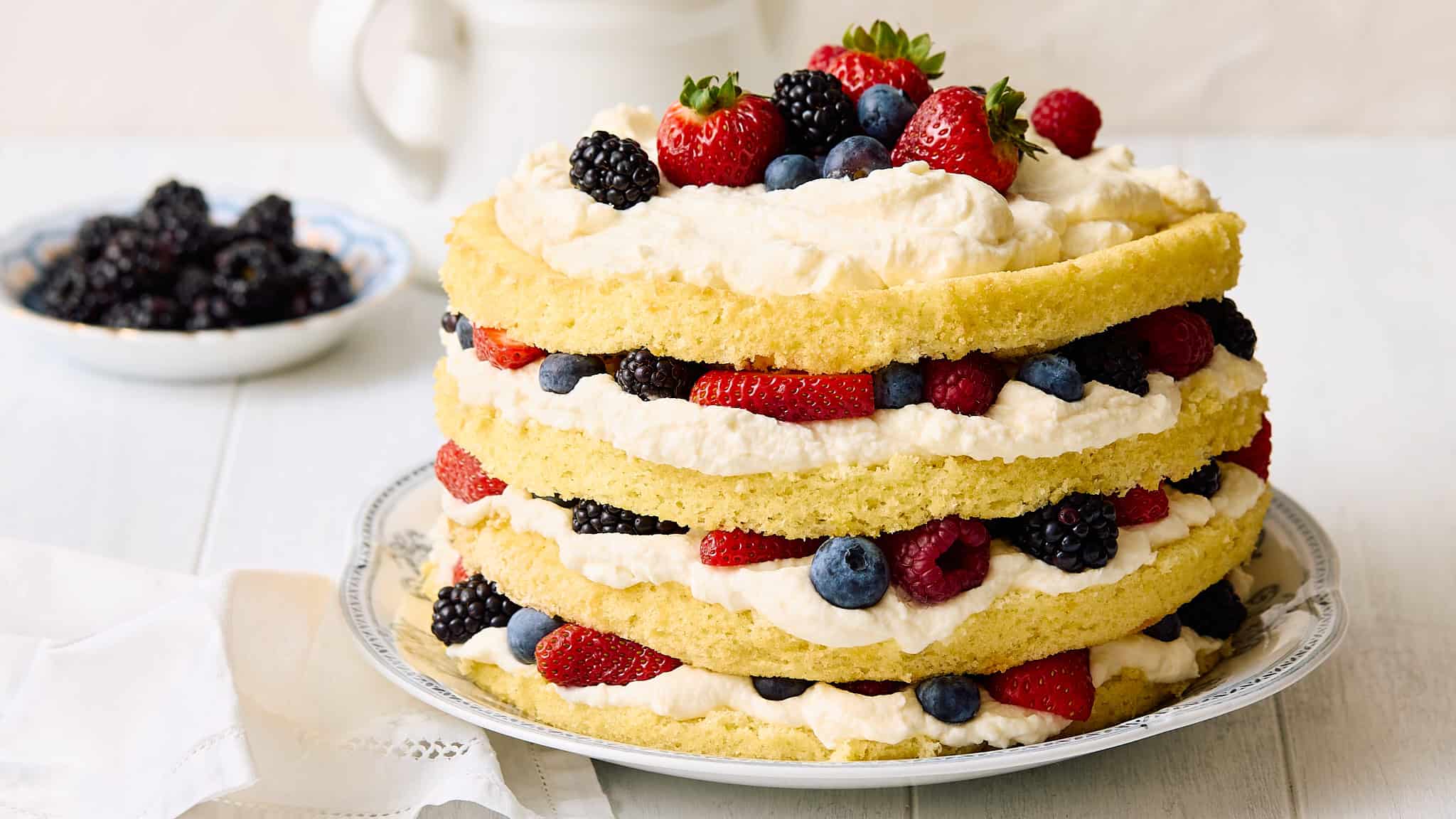 Fresh berry layered sponge cake with whipped cream and assorted berries on top and in between layers, served on a decorative plate for a delightful dessert.
