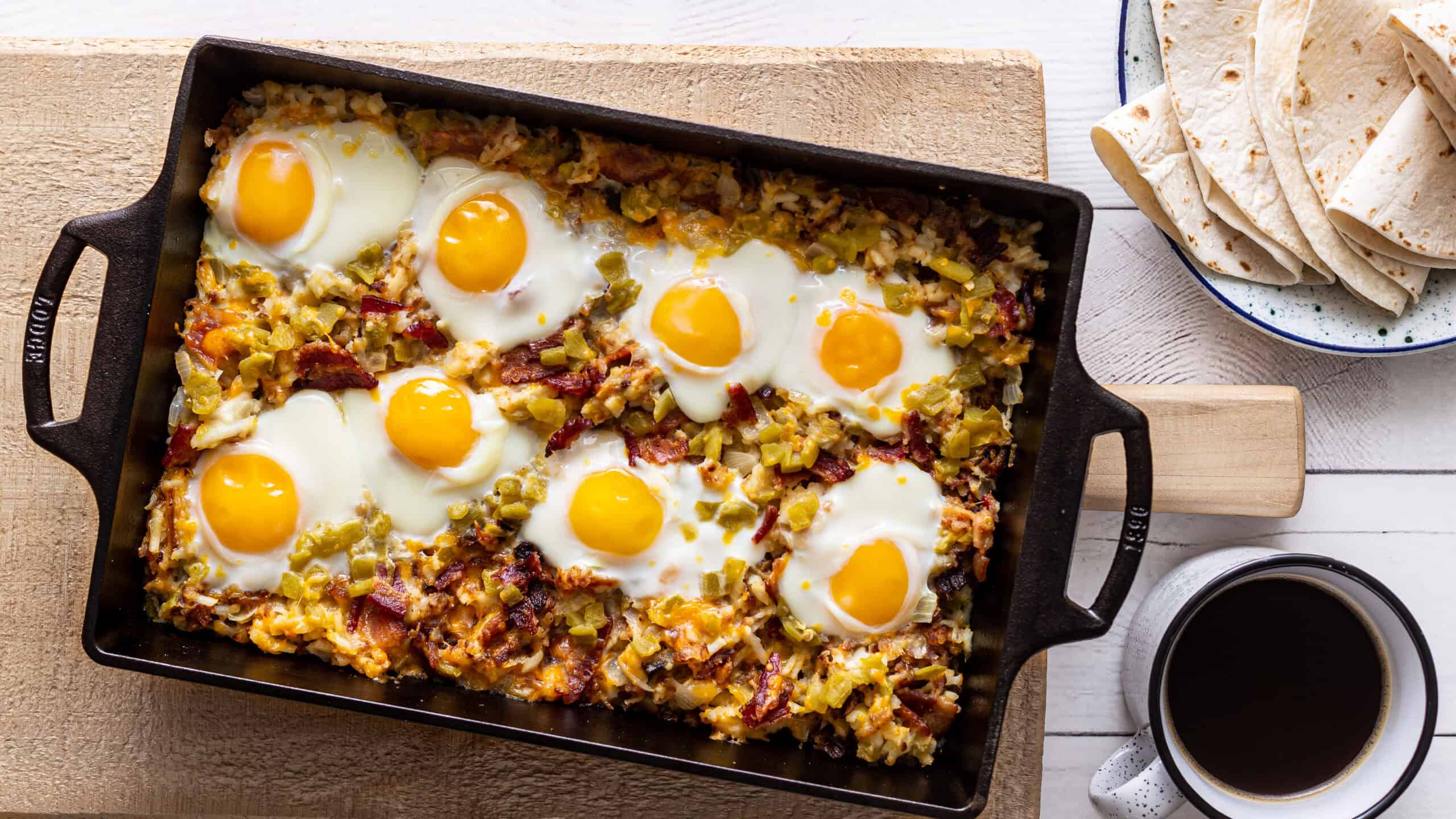 Savory breakfast casserole with baked eggs, bacon, and vegetables in a black baking dish, served with flour tortillas and coffee on a wooden table.