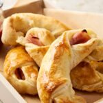 Golden baked ham and cheese croissants with flaky crust on a baking tray, served with a side of berry jam, perfect for breakfast or brunch.