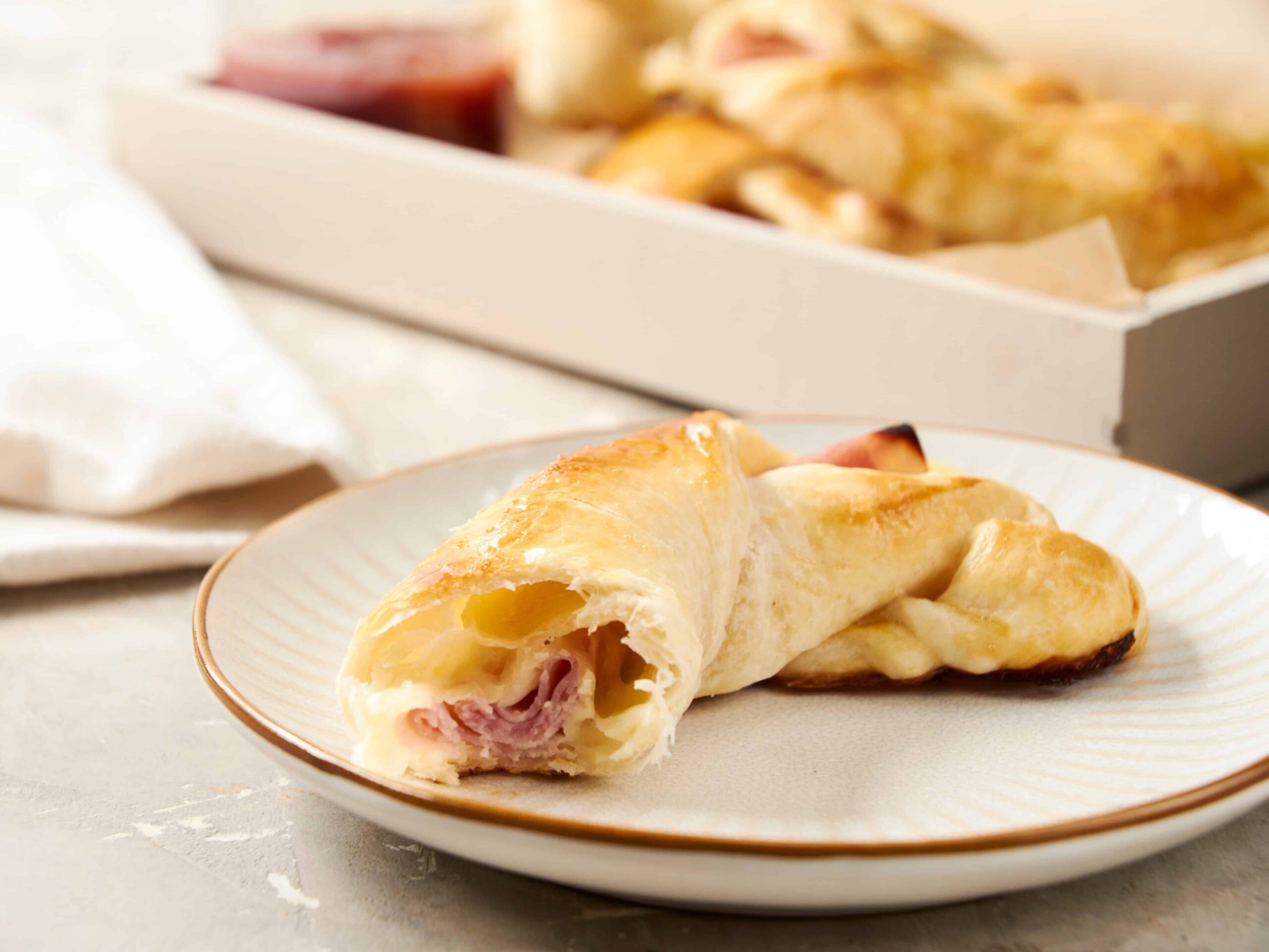Golden cheese and ham stuffed croissant on a white plate, with a baked pastry in the background, served as a delicious breakfast or brunch option.