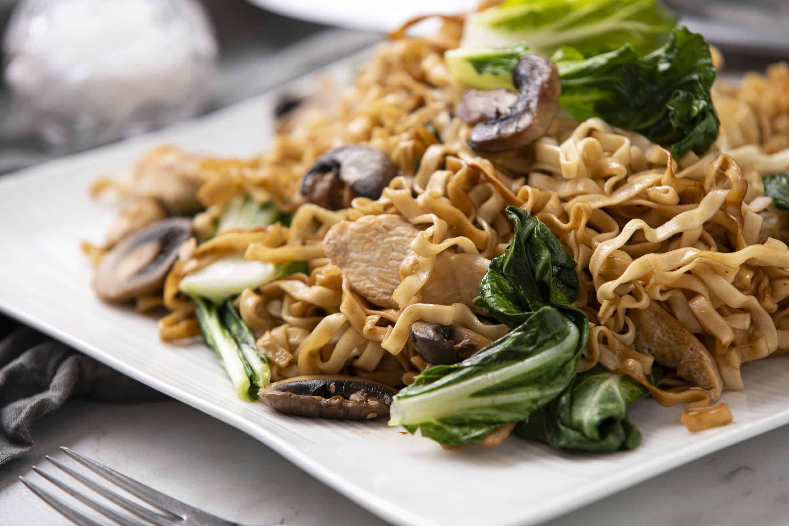 Fragrant stir-fried noodles with vegetables, mushrooms, and chicken served on a white plate, showcasing an Asian-inspired recipe perfect for quick and delicious meals.