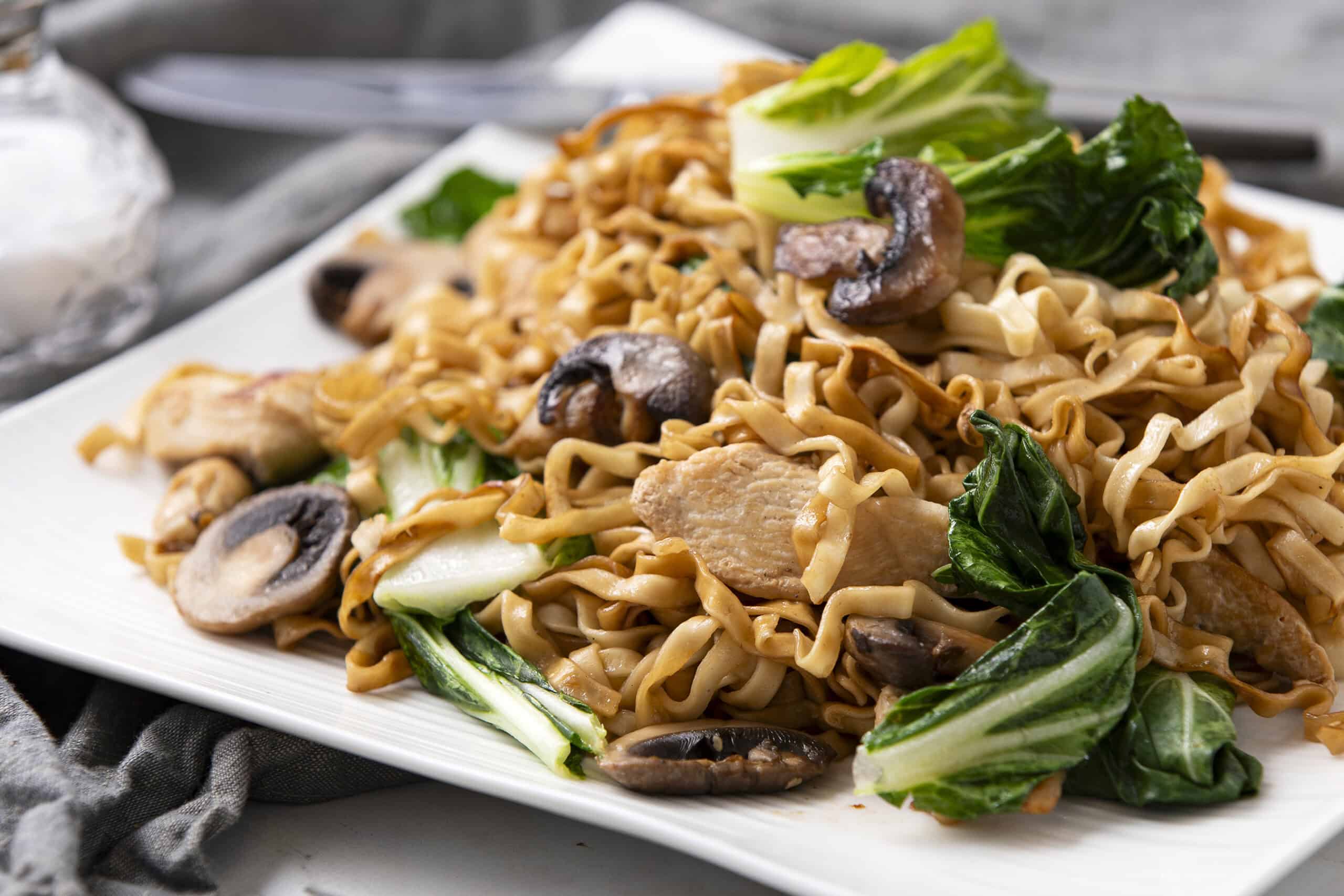 Sauteed chicken and vegetable stir-fry with noodles, mushrooms, and fresh greens on a white plate, served with soy sauce. Perfect for quick, healthy Asian-inspired meals.