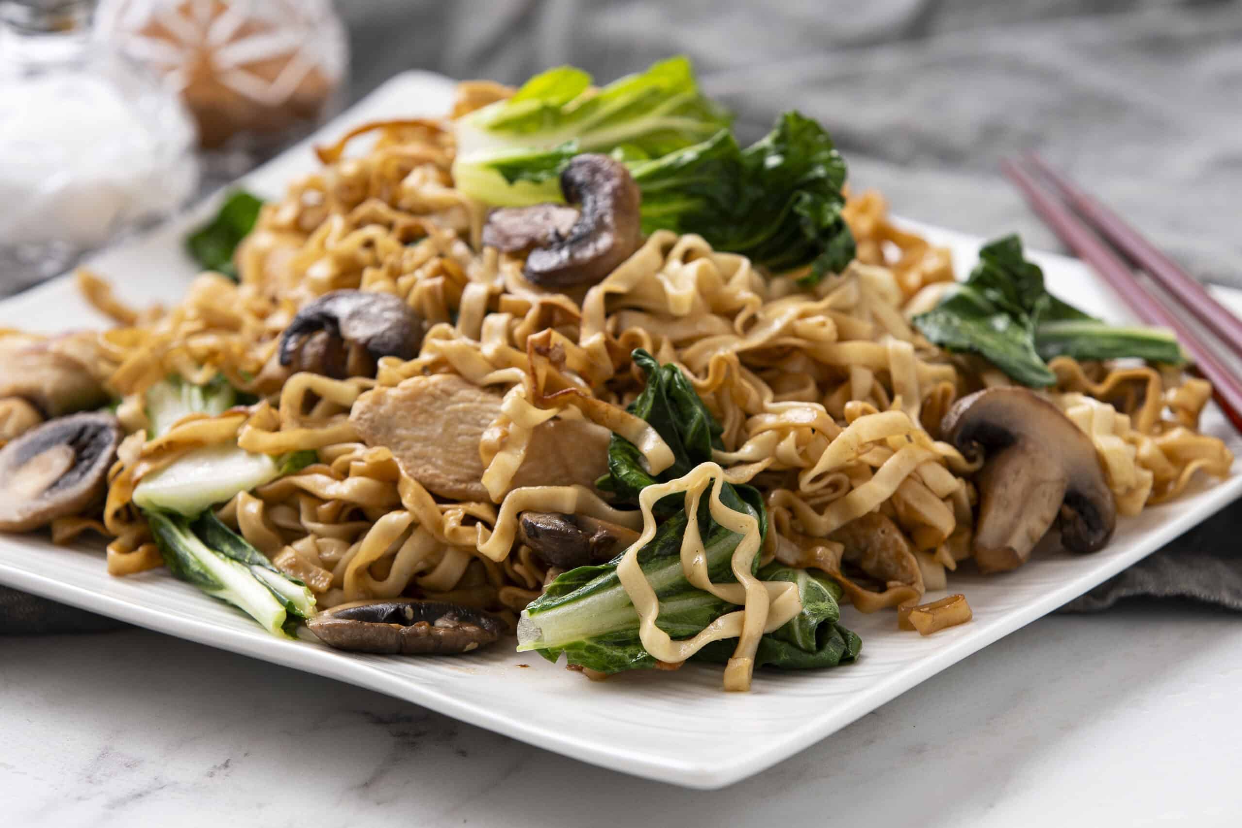 Stir-fried noodles with vegetables and mushrooms on a white rectangular plate, featuring bok choy and sliced chicken for a healthy Asian-inspired dish.