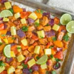 Freshly baked chicken and vegetable casserole topped with pineapple chunks, cilantro, and lime slices on a baking sheet. Colorful bell peppers, sweet potatoes, and purple onions create a vibrant, healthy dish.