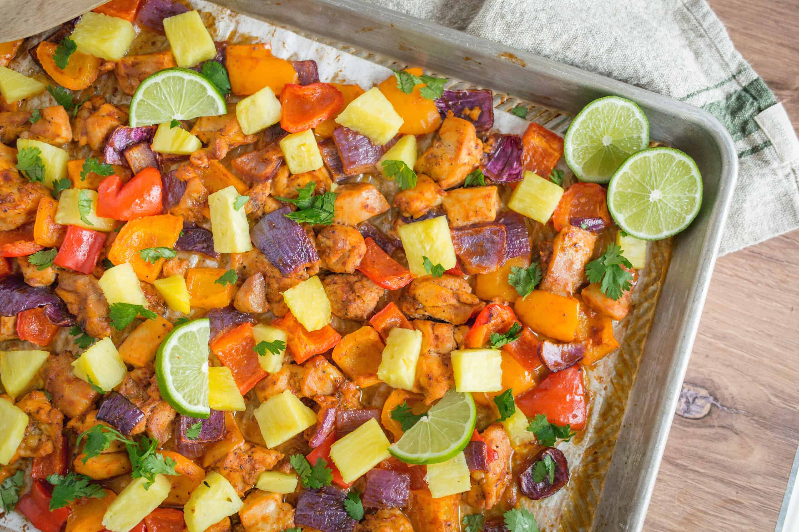 Freshly baked chicken and vegetable casserole topped with pineapple chunks, cilantro, and lime slices on a baking sheet. Colorful bell peppers, sweet potatoes, and purple onions create a vibrant, healthy dish.