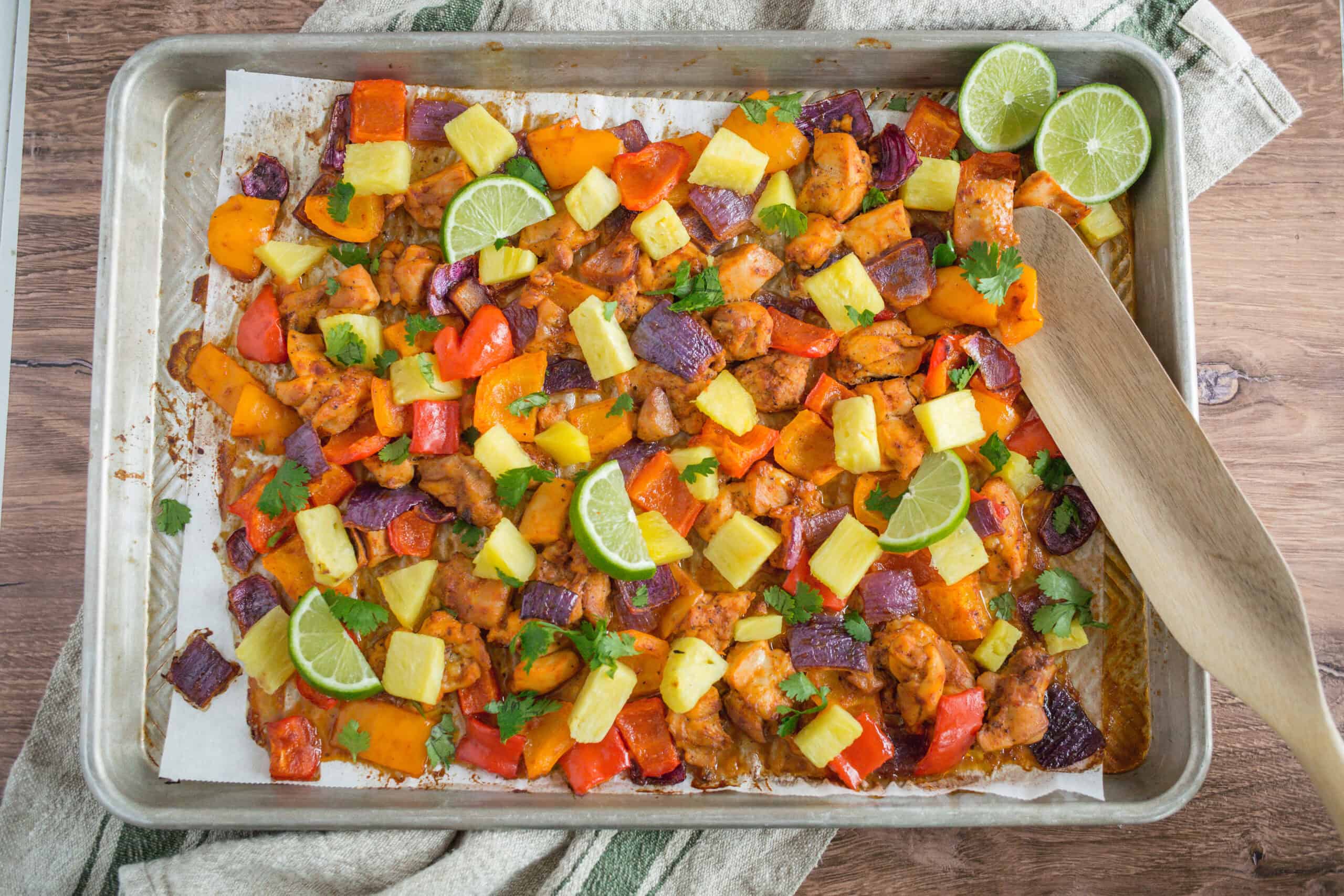 Roasted chicken and colorful vegetable sheet tray dinner with pineapple chunks, lime wedges, and cilantro garnish on a baking sheet.