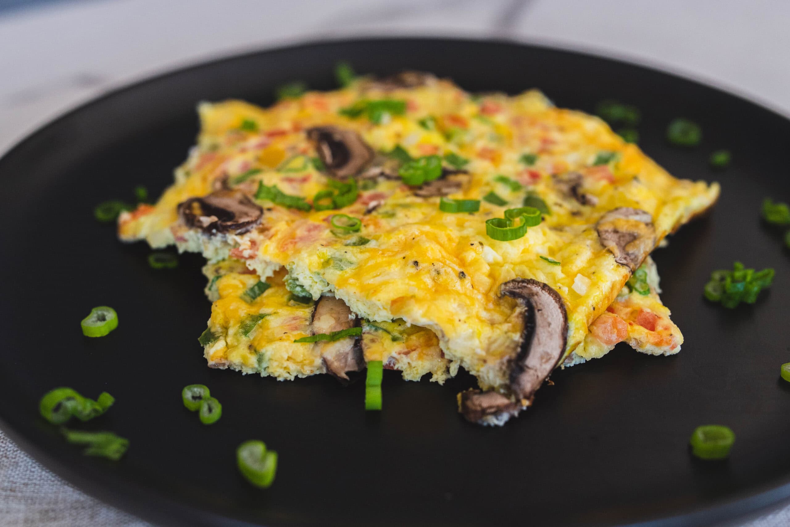 Fluffy mushroom and vegetable omelette garnished with chopped green onions on a black plate. Perfect for breakfast, brunch, or any meal, this delicious dish combines eggs, fresh vegetables, and savory mushrooms.