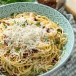 Creamy spaghetti carbonara with grated cheese and fresh herbs in a turquoise bowl. Fresh garlic, Parmesan cheese block, and dried chili flakes on a checkered cloth nearby. Perfect for a flavorful Italian dinner.