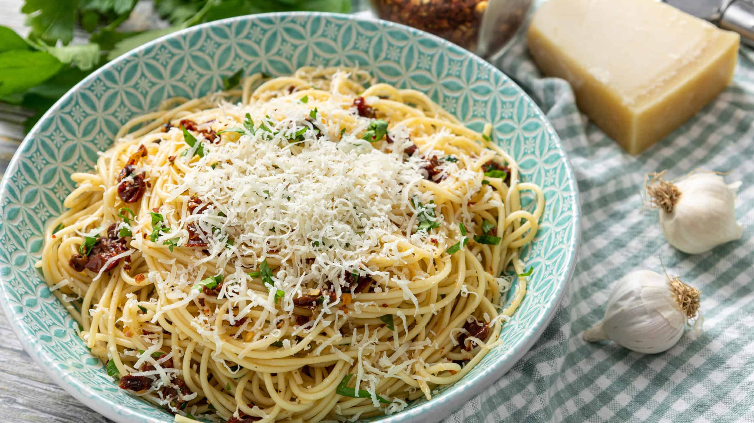 Creamy spaghetti carbonara with grated cheese and fresh herbs in a turquoise bowl. Fresh garlic, Parmesan cheese block, and dried chili flakes on a checkered cloth nearby. Perfect for a flavorful Italian dinner.