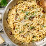 Creamy Parmesan spaghetti with sun-dried tomatoes and fresh herbs in a stainless steel skillet on rustic wooden background.