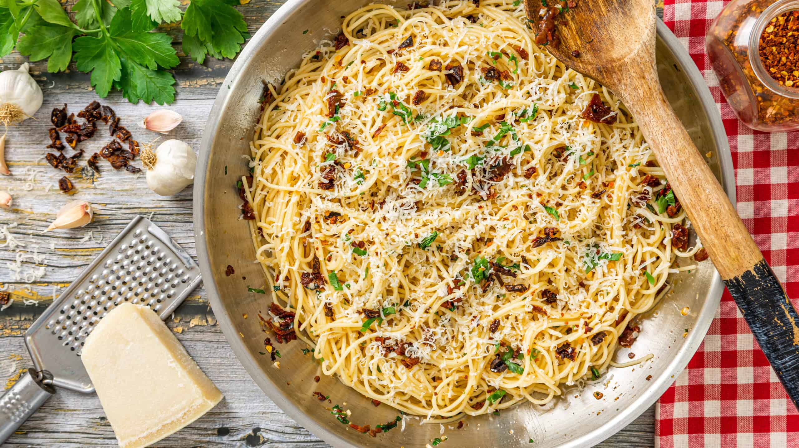 Creamy Parmesan spaghetti with sun-dried tomatoes and fresh herbs in a stainless steel skillet on rustic wooden background.