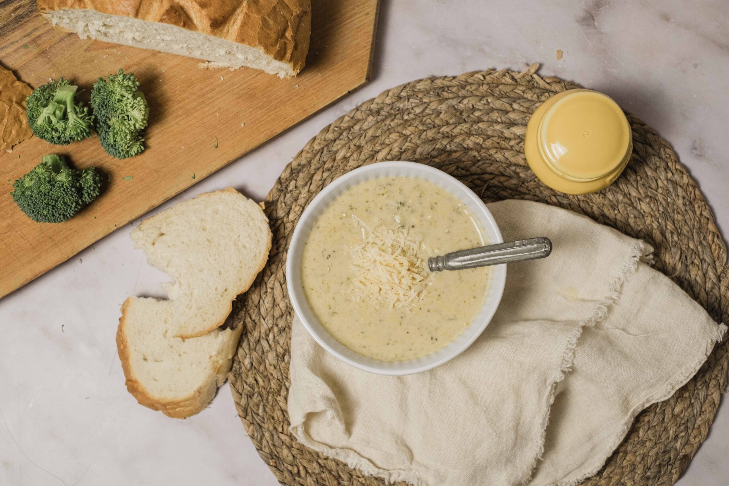 Creamy broccoli cheese soup served with shredded cheese, fresh broccoli florets, and sliced bread on a rustic woven placemat. Perfect for comfort food recipes and quick homemade soup ideas.
