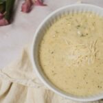 Creamy cauliflower cheese soup in a white bowl topped with shredded cheese, served on a neutral background with pink flowers and a linen cloth, perfect for comfort food recipes.