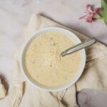 Creamy herb cheese dip in a white bowl with shredded cheese on top, served with fresh bread slices on a rustic cloth and pink lilies for garnish, perfect for appetizer or snack.