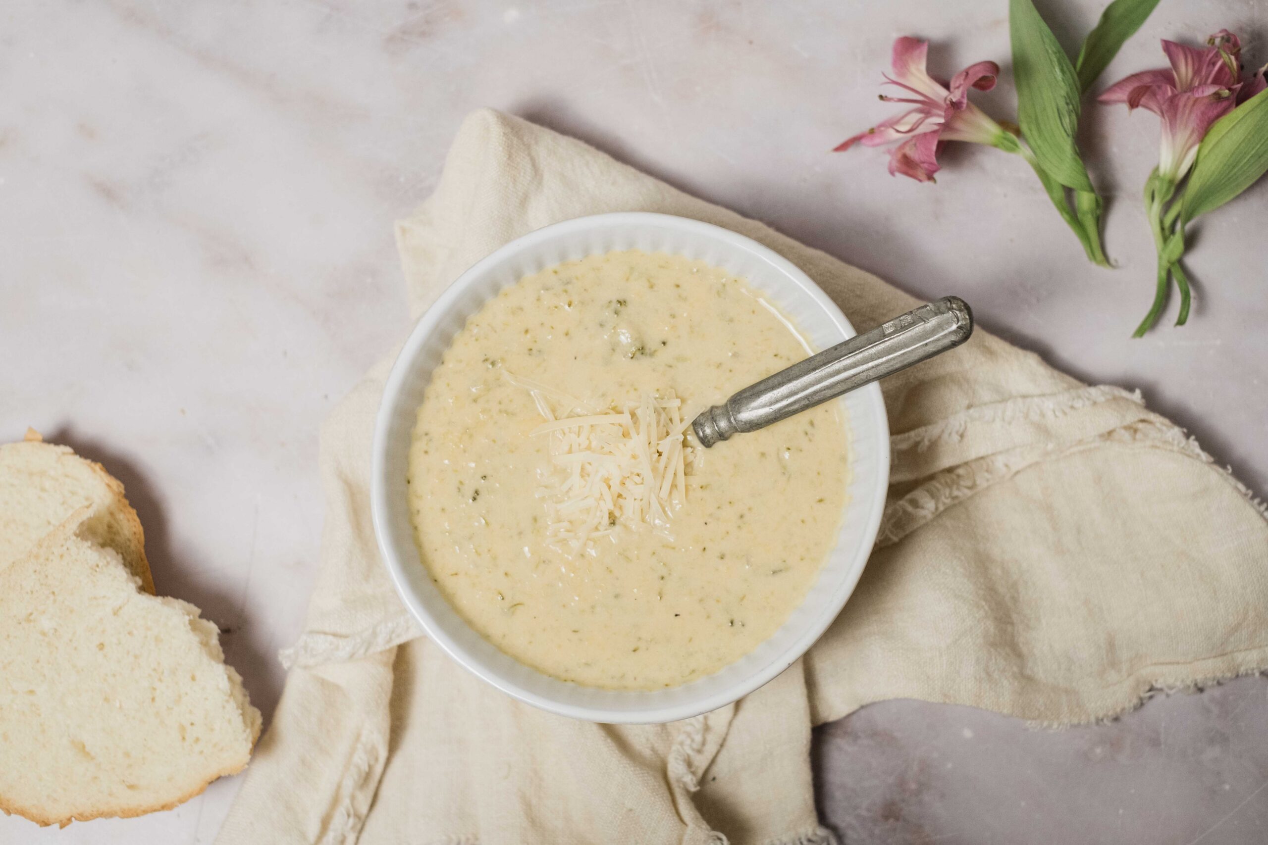 Creamy herb cheese dip in a white bowl with shredded cheese on top, served with fresh bread slices on a rustic cloth and pink lilies for garnish, perfect for appetizer or snack.