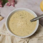 Creamy herb cheese soup in a white bowl garnished with shredded cheese, served with a slice of bread, yellow mustard bottle, and pink flowers in the background. Perfect for cozy meals and comfort food.