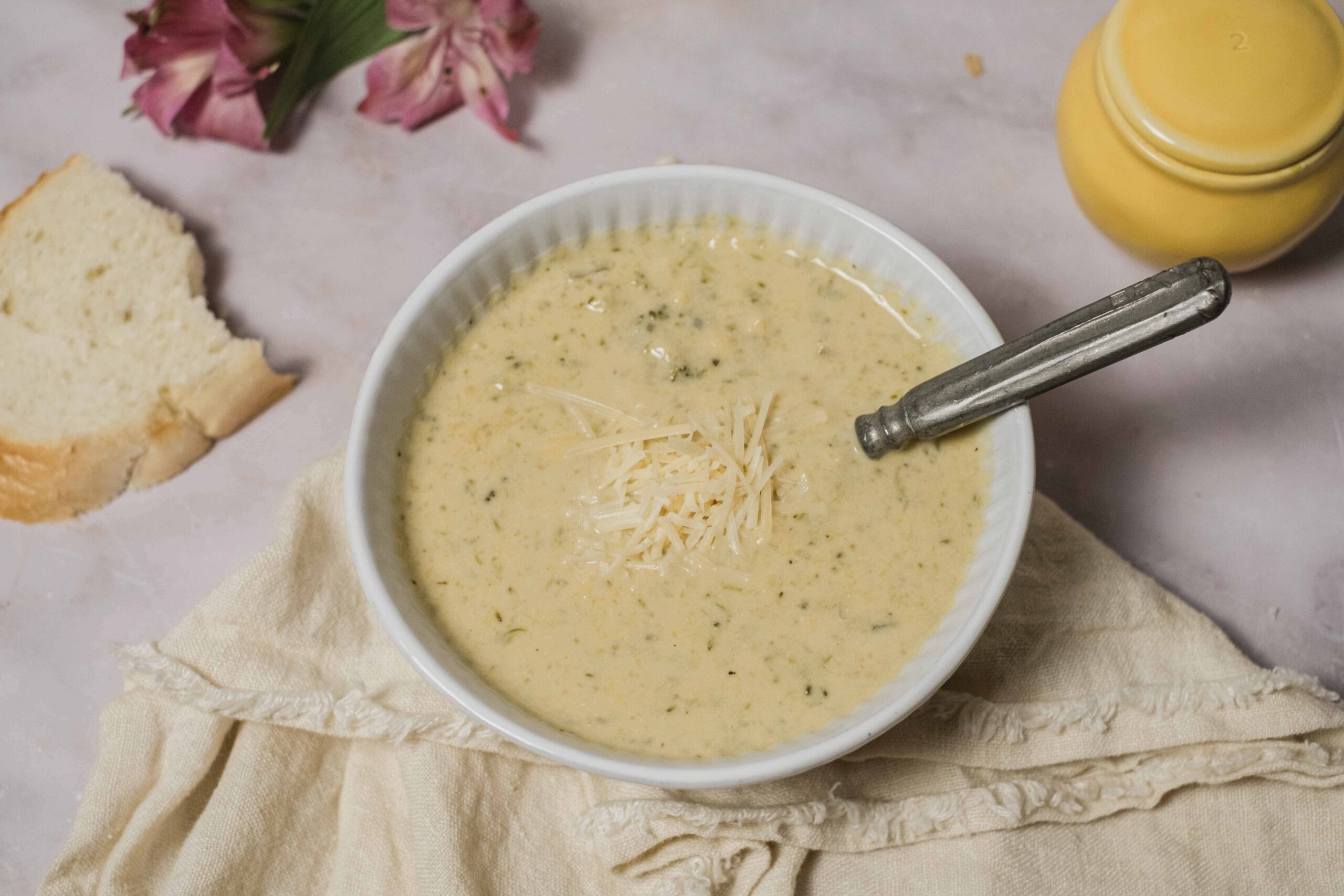 Creamy herb cheese soup in a white bowl garnished with shredded cheese, served with a slice of bread, yellow mustard bottle, and pink flowers in the background. Perfect for cozy meals and comfort food.