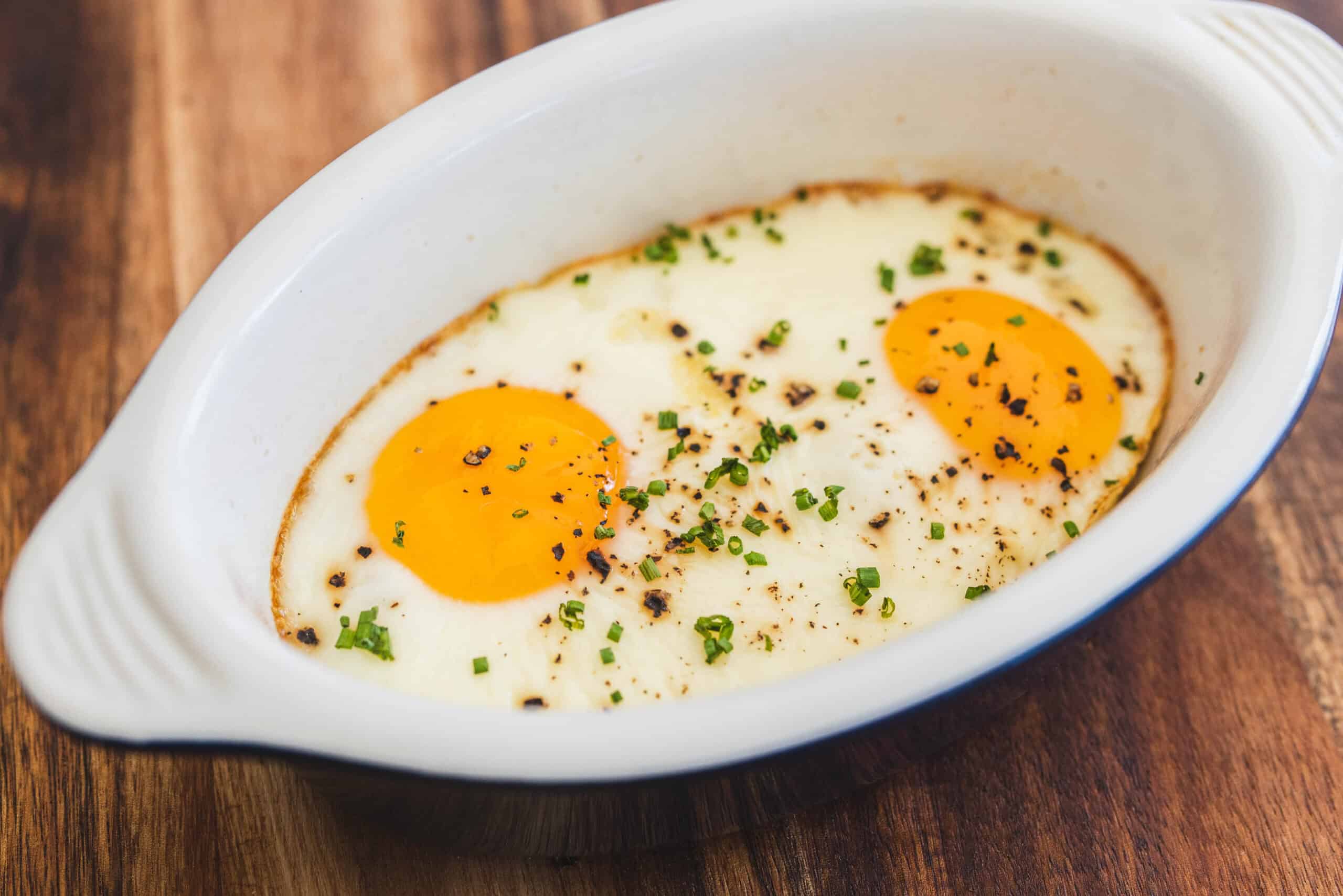 Creamy baked eggs with herbs and black pepper in a white oval dish on a wooden table.
