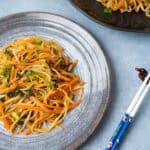 Stir-fried vegetable noodles with carrots, green onions, and leafy greens on a rustic plate, served with chopsticks on a blue surface, perfect for healthy Asian-inspired recipes.
