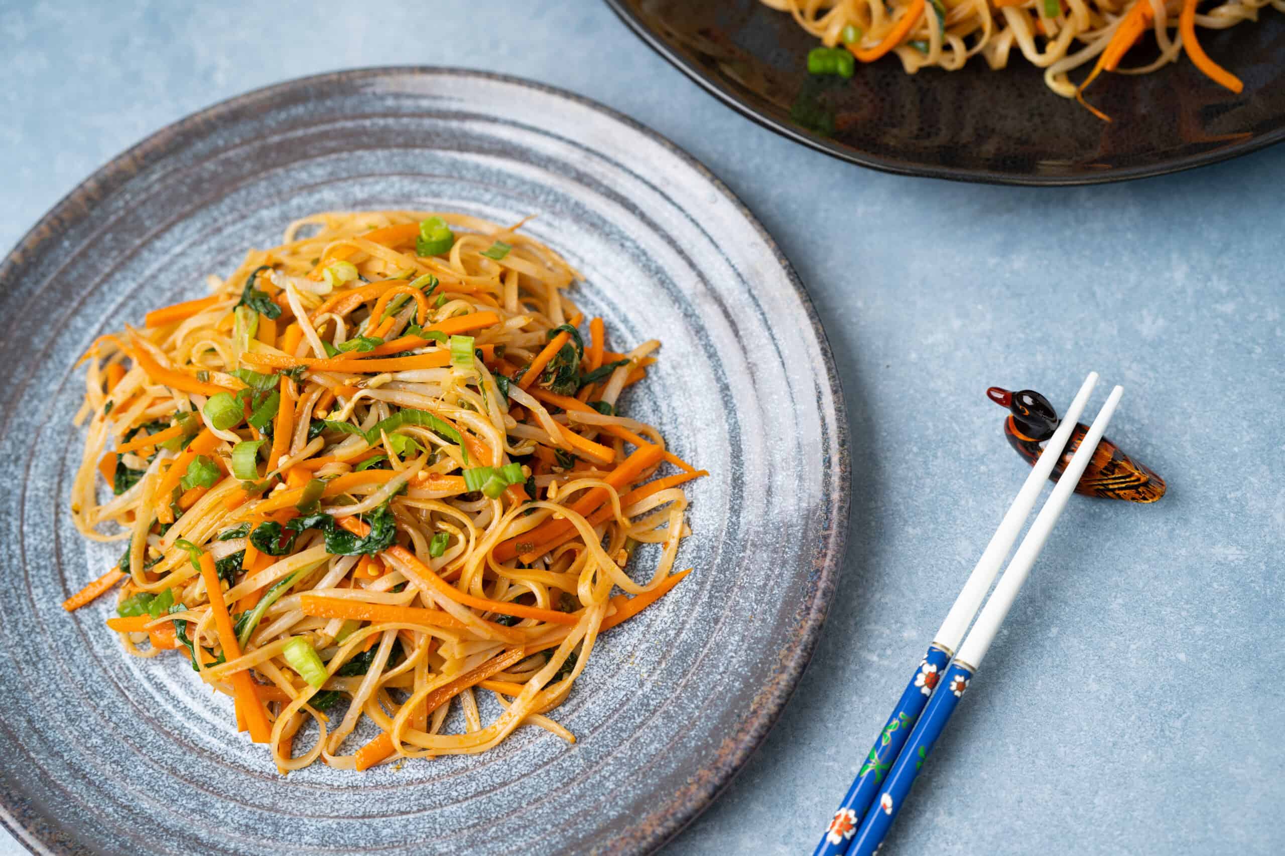 Stir-fried vegetable noodles with carrots, green onions, and leafy greens on a rustic plate, served with chopsticks on a blue surface, perfect for healthy Asian-inspired recipes.