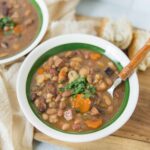 Comforting bean and vegetable soup in a white bowl with green rim, served with fresh herbs and bread on a wooden cutting board, perfect for healthy, hearty meals.