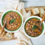 Hearty homemade bean and meat chili served in green-rimmed bowls with fresh bread, garnished with herbs, on a wooden board for a comforting meal.