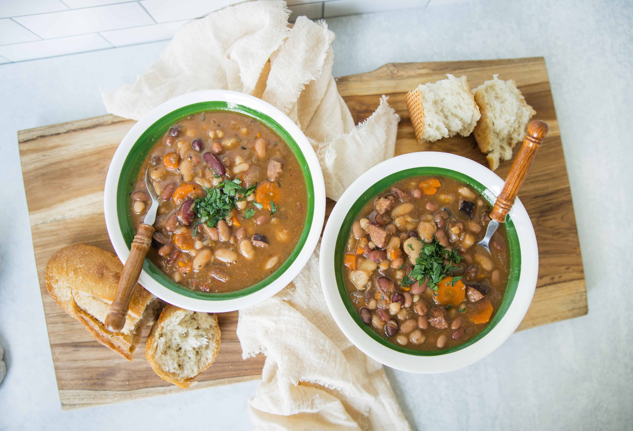 Hearty homemade bean and meat chili served in green-rimmed bowls with fresh bread, garnished with herbs, on a wooden board for a comforting meal.