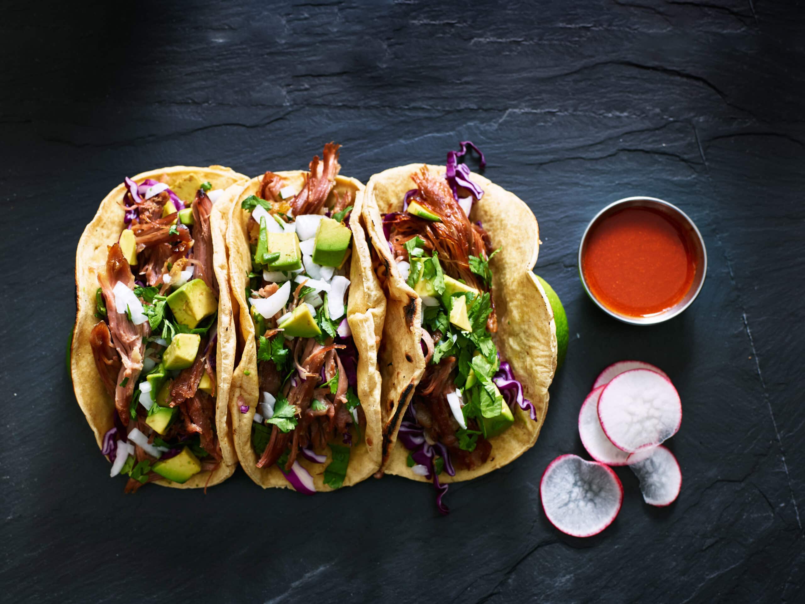 Succulent carne asada tacos with fresh avocado, cilantro, red cabbage, and chopped onions in soft corn tortillas, served with radish slices, a side of red salsa, and lime wedges on a dark slate background.