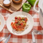 Savory stew served over rice on a plate with fresh green bell peppers and ingredients in the background.