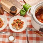 Savory homemade chicken stew served over white rice, cooked in a slow cooker with vegetables and spices. Fresh green bell peppers, salt, pepper, and cooking utensils are visible on the checkered tablecloth.