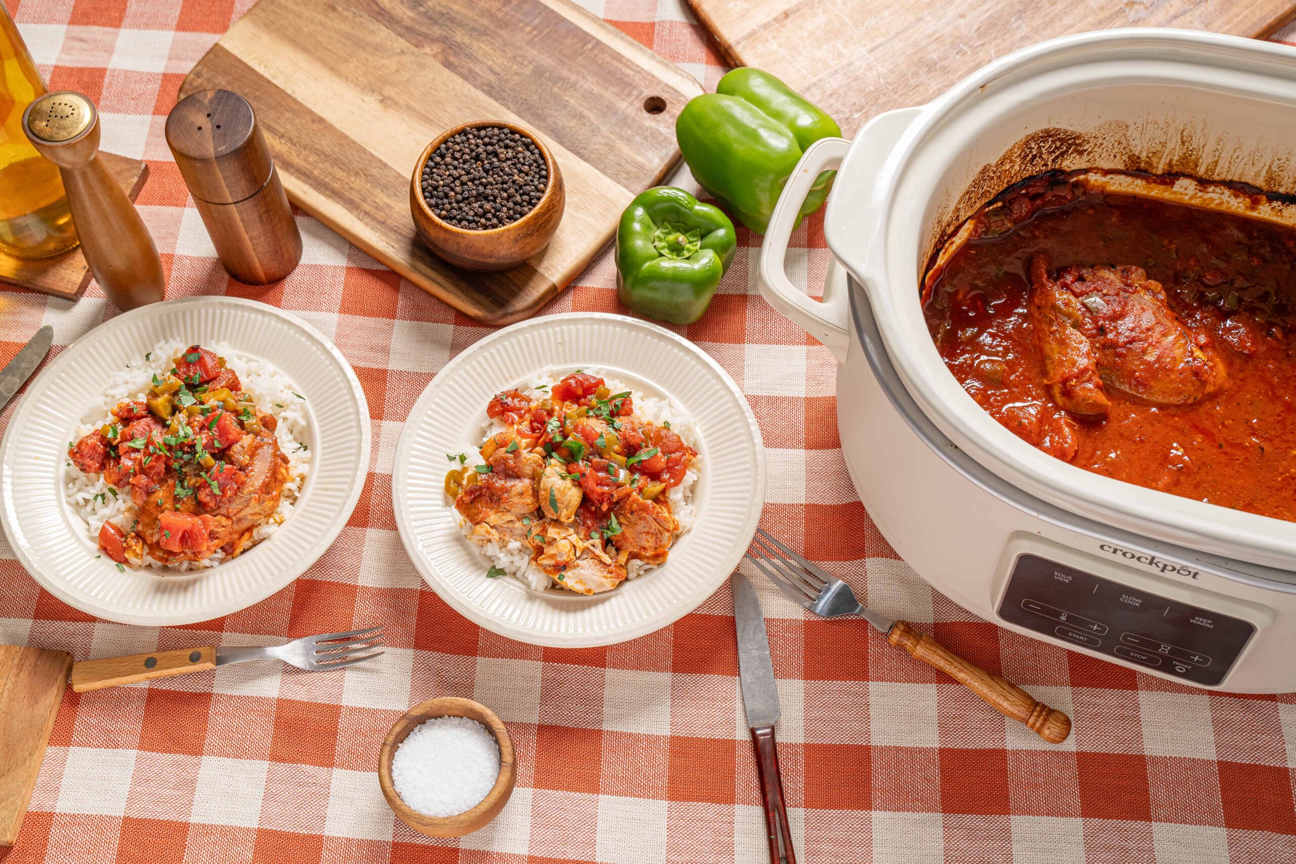 Savory homemade chicken stew served over white rice, cooked in a slow cooker with vegetables and spices. Fresh green bell peppers, salt, pepper, and cooking utensils are visible on the checkered tablecloth.